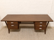 Load image into Gallery viewer, Mid-century modern wooden writing desk with a dark brown finish, featuring two drawers on each side with silver handles, set against a white brick wall and beige carpet.