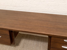 Load image into Gallery viewer, A dark brown wooden writing desk with drawers on both sides, shown against a white brick wall and textured beige carpet.
