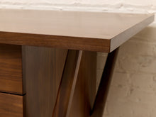 Load image into Gallery viewer, Close-up of a mid-century modern wooden desk corner with a warm brown walnut finish, showing the desktop edge, drawer front, and angled metal legs against a white brick wall.