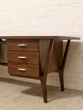 Load image into Gallery viewer, Mid-century modern wooden writing desk with three drawers and angled legs, shown against a white brick wall and beige carpet.