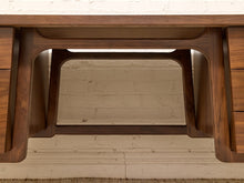 Load image into Gallery viewer, Close-up of a dark walnut wood writing desk showing the apron, legs, and drawer fronts against a white brick wall background.