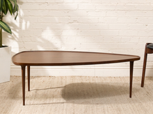 Load image into Gallery viewer, A mid-century modern wooden coffee table with a rounded triangular top and tapered legs, shown in a styled room setting against a white brick wall.