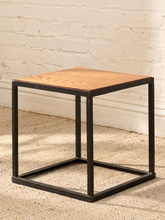 Load image into Gallery viewer, A square side table with a light brown wood grain top and a black metal frame, set on a textured beige rug against a white brick wall.