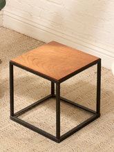 Load image into Gallery viewer, A square side table with a warm medium-brown wood grain top and a black metal cube frame base, set on a textured beige carpet against a white brick wall.