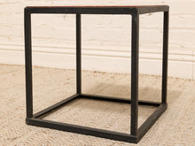 Load image into Gallery viewer, A cube-shaped side table with a black metal frame and a reddish-brown wooden top, set on a beige textured rug against a white brick wall.