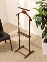 Load image into Gallery viewer, A mid-century modern wooden coat rack with a hanger and shelf, shown in a styled room setting with a white brick wall, beige carpet, a black chair, and a potted plant.