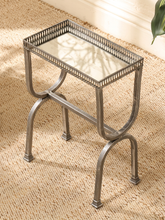 Load image into Gallery viewer, A small rectangular side table with a mirrored glass top and a decorative silver metal frame, shown on a woven beige rug in a styled interior setting.