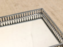 Load image into Gallery viewer, Close-up of a decorative rectangular mirror with an ornate silver-toned metal frame featuring a repeating balustrade pattern, resting on a beige textured surface.