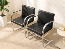 Load image into Gallery viewer, Two modern black leather armchairs with curved chrome metal frames, shown in a bright room with a white brick wall, beige carpet, and a potted plant.