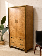 Load image into Gallery viewer, A tall vintage-style wooden cabinet with a burl wood grain finish and brass trim, featuring two doors with double handles and two drawers at the bottom, set against a white brick wall.
