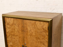 Load image into Gallery viewer, A mid-century modern wooden cabinet featuring a rich burl wood grain door panel and polished brass trim, set against a white brick wall background.