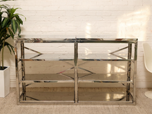 Load image into Gallery viewer, A modern rectangular console table with a polished chrome frame, mirrored glass shelves, and geometric metal corner accents, set against a white brick wall with a potted plant on the left.