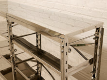Load image into Gallery viewer, A polished stainless steel industrial shelving unit with a thick top work surface and multiple adjustable metal shelves, shown against a white brick wall background.