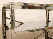 Load image into Gallery viewer, Close-up of a polished stainless steel shelving unit with adjustable metal brackets, showing reflective surfaces and hardware against a white brick wall background.