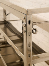 Load image into Gallery viewer, Close-up of a stainless steel shelving unit corner with a hanging hook and adjustment holes, photographed against a light brick wall.
