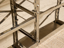 Load image into Gallery viewer, Close-up of a polished stainless steel industrial shelving system with adjustable uprights and horizontal beams, set against a light brick wall and textured beige floor.