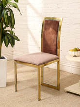 Load image into Gallery viewer, A modern dining chair with a curved gold metal frame, a brown velvet backrest, and a light beige upholstered seat cushion, set against a white brick wall with a potted plant and a small side table.