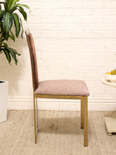 Load image into Gallery viewer, A modern dining chair with a light brown upholstered seat and backrest, supported by a gold-finished metal frame, set against a white brick wall background.