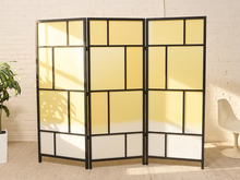 Load image into Gallery viewer, A three-panel folding room divider with a black metal frame and frosted glass panels in yellow, cream, and white, styled in a bright room with a white brick wall, a potted plant, and a chair.