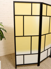 Load image into Gallery viewer, A three-panel folding room divider with a black metal frame and frosted panels that fade from pale yellow at the top to white at the bottom, set against a white brick wall and beige carpet.