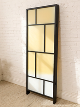 Load image into Gallery viewer, A tall, narrow black metal screen or room divider with frosted glass panels in a geometric grid pattern, leaning against a white brick wall on a beige carpet.