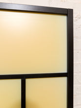 Load image into Gallery viewer, Close-up of a modern black-framed wall divider or room divider with frosted cream panels, set against a white brick wall.
