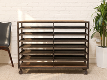 Load image into Gallery viewer, A dark brown industrial-style rolling storage cabinet with multiple horizontal shelves and caster wheels, set against a white brick wall with a potted plant and chair partially visible.