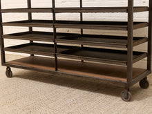 Load image into Gallery viewer, A dark metal rolling storage rack with multiple tiers and caster wheels, shown on a textured beige carpet against a white brick wall.