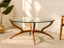 Load image into Gallery viewer, A mid-century modern round glass-top coffee table with a sculptural wooden base, set on a textured beige rug against a white brick wall.