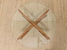 Load image into Gallery viewer, A round clear glass table top with an X-shaped wooden base, photographed from directly above on a textured beige woven rug.