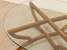Load image into Gallery viewer, Close-up of a round glass-top table with a sculptural wooden base made of interlocking angular wood beams, shown on a woven beige rug.