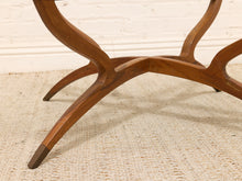 Load image into Gallery viewer, Close-up of a mid-century modern wooden table base with curved legs and tapered feet, resting on a textured beige rug against a white brick wall.