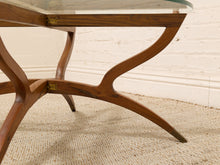 Load image into Gallery viewer, A close-up of a mid-century modern wooden table base with curved legs and a clear glass tabletop, set on a textured beige rug against a white brick wall.