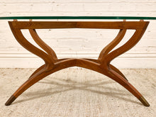 Load image into Gallery viewer, A mid-century modern wooden table base with a curved, sculptural X-shaped design and a clear glass top, set against a white brick wall and textured beige rug.