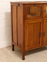 Load image into Gallery viewer, A vintage wooden cabinet with a warm medium-brown finish, featuring a glass-front upper cabinet door, solid wood lower doors, brass-style hardware, and turned legs, set against a white brick wall and beige rug.