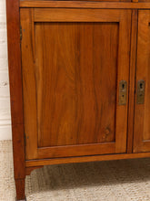Load image into Gallery viewer, Close-up of a wooden cabinet door with warm medium-brown wood grain, brass-style keyhole hardware, and part of the cabinet frame visible against a white brick wall and woven rug.