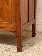 Load image into Gallery viewer, Close-up of a warm medium-brown wooden cabinet or sideboard showing the corner joint, paneling, and turned legs on a woven rug against a white brick wall.