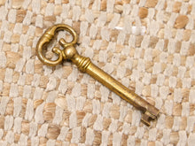 Load image into Gallery viewer, An antique brass key with an ornate bow and a weathered finish, resting diagonally on a woven beige and white textured mat.