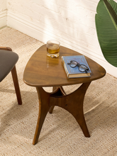 Load image into Gallery viewer, A triangular wooden side table with a sculptural curved base, styled in a home setting with a glass of whiskey, a blue book, and eyeglasses on top, set on a textured beige rug against a white brick wall.