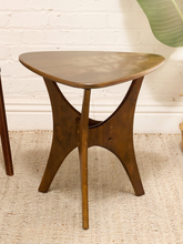 Load image into Gallery viewer, A mid-century modern wooden end table with a triangular top and three splayed legs, shown on a textured beige rug against a white brick wall.