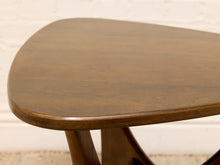 Load image into Gallery viewer, Close-up of a mid-century modern wooden stool with a rounded triangular seat and curved legs, set against a white brick wall.