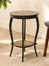 Load image into Gallery viewer, A small round accent side table with a distressed black metal frame and a light brown wood top and lower shelf, set on a beige textured rug against a white brick wall.