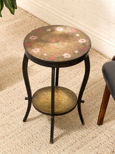Load image into Gallery viewer, A small round metal side table with a floral pattern on the top surface and a lower shelf, shown in a home setting on a textured rug next to a chair.