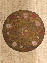 Load image into Gallery viewer, A round decorative mirror with an intricate gold-toned floral pattern and pink and red blossoms, set against a textured beige woven background.