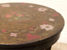 Load image into Gallery viewer, Close-up of a round wooden stool or table top with a dark black lacquered base and hand-painted floral motifs in pink, white, and muted green over a brown wood grain.