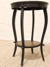 Load image into Gallery viewer, A vintage black wooden side table with curved legs, a top tier, and a lower shelf, set against a white brick wall and beige carpet.