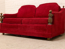 Load image into Gallery viewer, A vintage red upholstered loveseat with dark wood turned post arms and brass caster wheels, set against a white brick wall and light carpet.