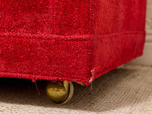 Load image into Gallery viewer, Close-up of the corner of a red upholstered piece of furniture with a brass caster wheel, resting on a beige woven rug.