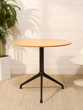 Load image into Gallery viewer, Round blonde wood dining table with simple circular top and sculptural black three-leg tripod base.
