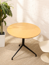 Load image into Gallery viewer, A round light wood table with a black metal tripod base, set in a room corner with a white brick wall, beige carpet, a potted plant, and a partial white chair.